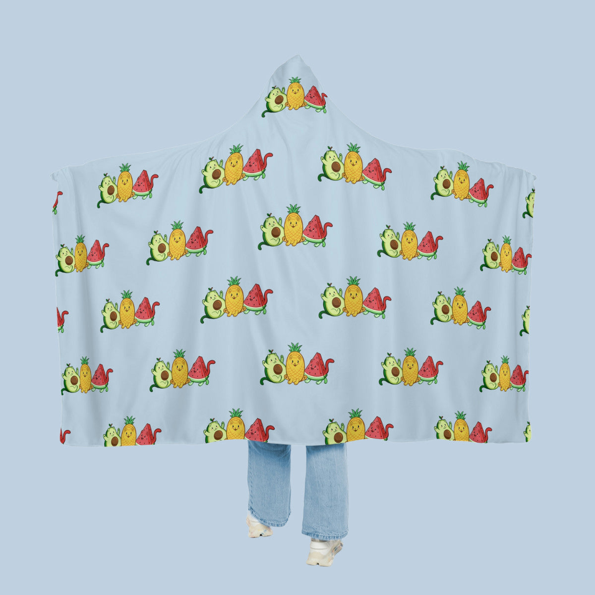 Happy woman wearing the light blue Fruitkittens⢠logo hooded microfiber fleece blanket like a cape with arms stretched out seen from behind so that the full pattern is visible. Fruitkittens⢠are fruits drawn as cute cats. The logo features Pawocadoâ˘, Pawnapple⢠and Watermewlonâ˘