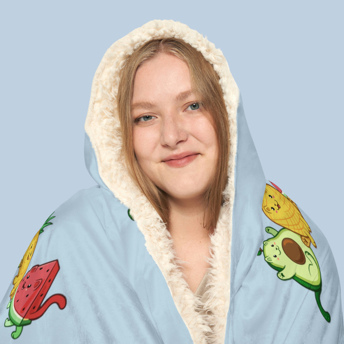 Happy woman wearing the light blue Fruitkittens⢠logo hooded sherpa blanket face zoomed in. Fruitkittens⢠are fruits drawn as cute cats. The logo features Pawocadoâ˘, Pawnapple⢠and Watermewlonâ˘