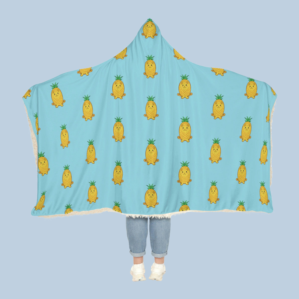 Happy woman wearing the light blue Pawnapple⢠hooded sherpa blanket like a cape with arms stretched out seen from behind so that the full pattern is visible. Pawnapple⢠is a pineapple drawn as a cute cat.