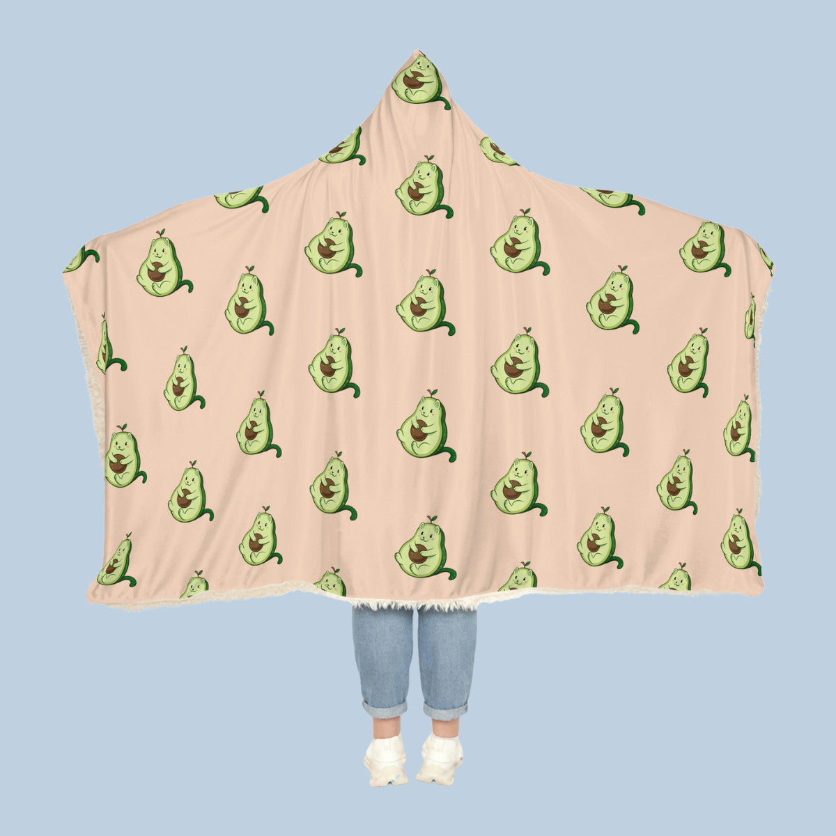 Happy woman wearing the beige Pawocado⢠hooded sherpa blanket like a cape with arms stretched out seen from behind so that the full pattern is visible. Pawocado⢠is an avocado drawn as a cute cat.