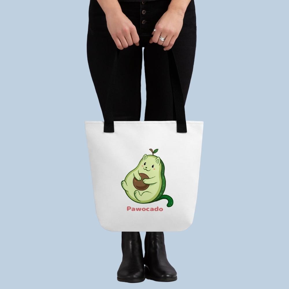 Person holding a white Tote Bag with Pawocado⢠print. Pawocado⢠is an avocado drawn as a cute cat.