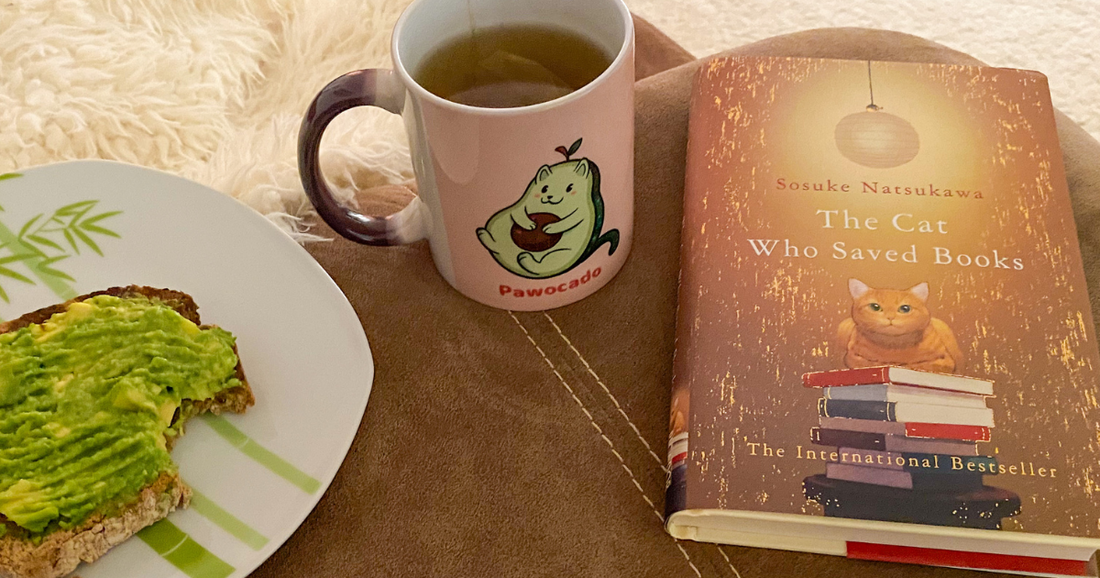 The adorable Pawocado™ Coffee Mug in a cozy setting with the book “The Cat That Saved Books” by Sosuke Natsukawa next to it. Pawocado™ is a cute cat drawn like an avocado.