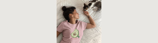 Woman wearing the pink Pawocado™ T-shirt snuggling in bed with a cat. Pawocado™ is an avocado drawn like an adorable kitten.