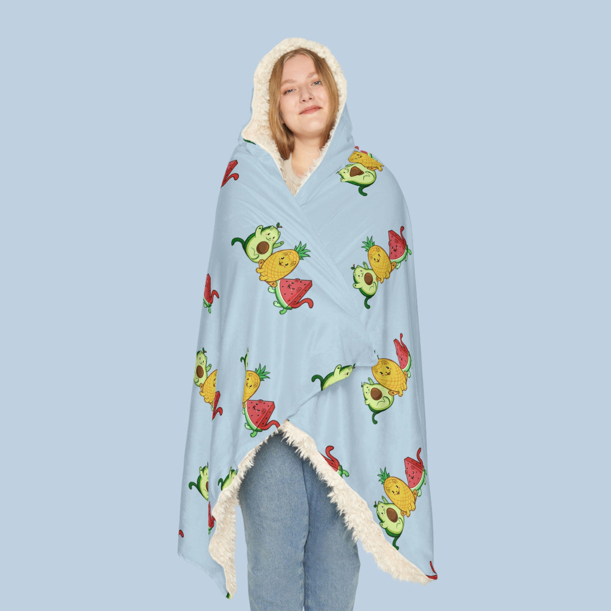 Happy woman wearing the light blue Fruitkittens™ logo hooded sherpa blanket seen from front. Fruitkittens™ are fruits drawn as cute cats. The logo features Pawocado™, Pawnapple™ and Watermewlon™