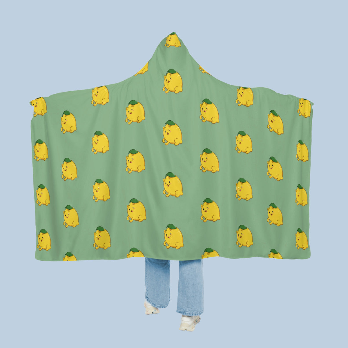 Happy woman wearing the light green Lemeow™ hooded microfiber fleece blanket like a cape with arms stretched out seen from behind so that the full pattern is visible. Lemeow™ is a lemon drawn as a cute cat.