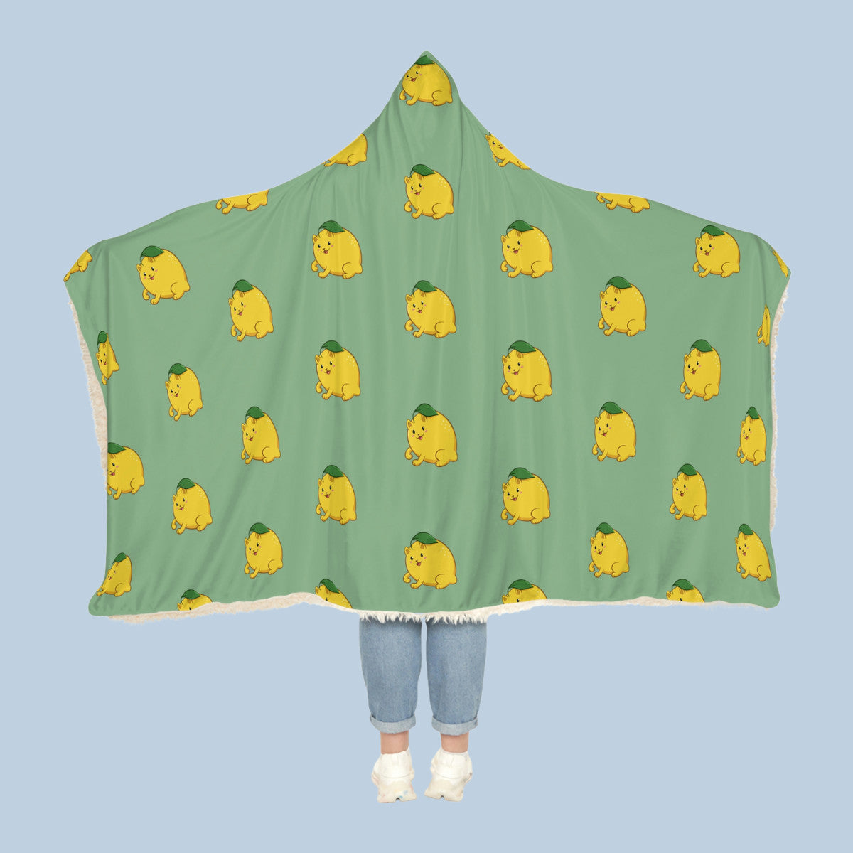 Happy woman wearing the light green Lemeow™ hooded sherpa blanket like a cape with arms stretched out seen from behind so that the full pattern is visible. Lemeow™ is a lemon drawn as a cute cat.