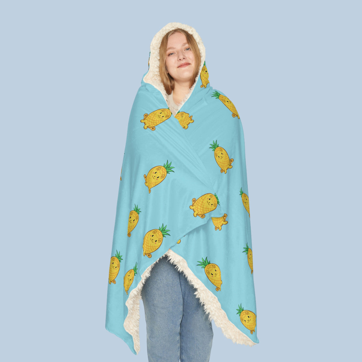 Happy woman wearing the light blue Pawnapple™ hooded sherpa blanket seen from front. Pawnapple™ is a pineapple drawn as a cute cat.